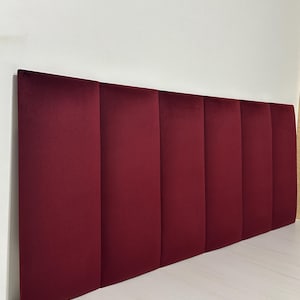 May include: A burgundy velvet headboard with a vertical panel design. The headboard is rectangular and leans against a white wall. The headboard is made of seven vertical panels.
