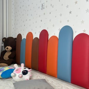 May include: A colorful headboard with a row of vertical, rounded panels in shades of brown, orange, red, and blue. A teddy bear and stuffed animals are visible on the bed. The wall behind the headboard is decorated with a star pattern.