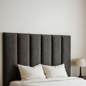 May include: A dark grey upholstered headboard with vertical panels. Two white pillows on a bed with a white duvet. A small lamp with a beige shade sits on a bedside table.