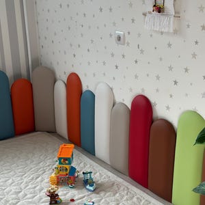 May include: A child's bedroom featuring a bed with a multi-coloured padded headboard. The headboard has vertical panels in blue, orange, white, grey, red, brown, and green. Toy building blocks are arranged on the bed.