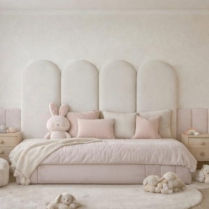 May include: A light pink and cream-colored bedroom with a large bed, plush toys, and a bunny-themed chair. The bed has a large headboard and is adorned with pillows and a blanket. A round rug and a small bookshelf complete the cozy space.