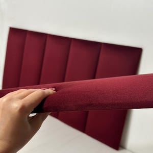 K&ouml;nnte beinhalten: Ein rechteckiges Kopfteil aus tiefburgunderfarbenem Samtstoff mit einer Reihe vertikaler, gepolsterter Paneele. Die Struktur ist aus einem seitlichen Winkel dargestellt, was die Dicke und die weiche Textur des Materials hervorhebt.