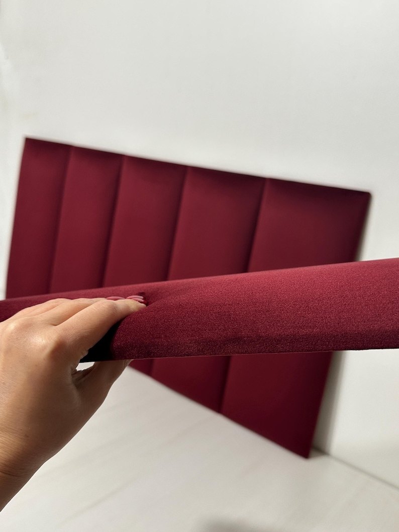 K&ouml;nnte beinhalten: Ein rechteckiges Kopfteil aus tiefburgunderfarbenem Samtstoff mit einer Reihe vertikaler, gepolsterter Paneele. Die Struktur ist aus einem seitlichen Winkel dargestellt, was die Dicke und die weiche Textur des Materials hervorhebt.