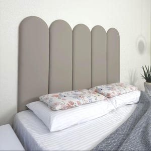 May include: A beige upholstered headboard with a curved top design. Two floral patterned pillows rest on a white bed with a striped comforter. A grey blanket and a potted plant are also visible.