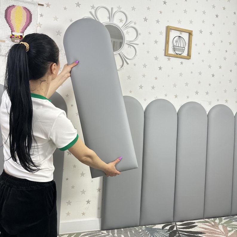 May include: A series of vertical, gray, padded wall panels with rounded tops. Each panel features a smooth, matte finish and a uniform, arched shape. The panels are arranged side-by-side to form a continuous, modular headboard-like structure.