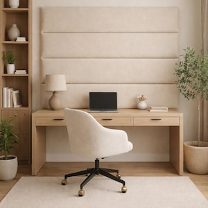 May include: A modern home office setup featuring a light wood desk with a laptop, lamp, and chair. A beige rug and a tall bookshelf with plants and decor complete the space. The wall behind the desk has a padded, beige panel.