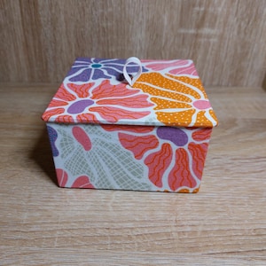 Può includere: Piccola scatola rettangolare in tessuto con coperchio. Decorata con un motivo floreale arancione, rosa, viola e bianco. Un anello di nastro bianco è attaccato al coperchio. Adatta per riporre oggetti o come elemento decorativo. Misure in cm.