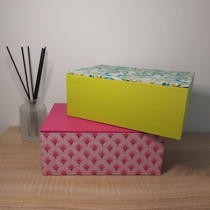 Può includere: Due scatole decorative impilate su una superficie di legno. La scatola inferiore è rosa con un motivo a ventaglio. La scatola superiore è verde lime con un coperchio fantasia con disegni floreali blu e verdi. È visibile un diffusore in vetro con canne nere.