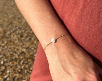 Puede incluir: Un delicado brazalete de cadena dorada con un solo dije de piedra blanca. El brazalete se muestra en la muñeca de una persona.