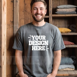 Może przedstawiać: Mężczyzna w szarej koszulce z nadrukiem "YOUR DESIGN HERE" na przodzie. Stoi przed drewnianą półką z poduszkami.