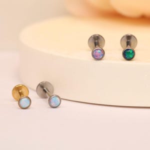 Puede incluir: Una colección de pequeños pendientes de botón con parte trasera plana y piedras redondas de ópalo iridiscente. Los pendientes son de varios colores, incluyendo oro, plata y tonos ópalo. Los pendientes se muestran sobre una superficie blanca.