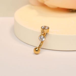 Puede incluir: Anillo de ombligo dorado con dos piedras de cristal transparentes. El anillo tiene una pequeña bola en la parte inferior.