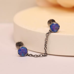 May include: A pair of silver-toned earrings connected by a delicate chain. Each earring features a round, blue opal stone. The earrings are displayed on a white surface with a soft, blurred background.