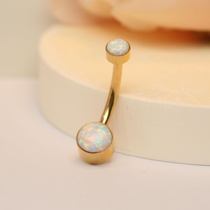 Puede incluir: Anillo de ombligo dorado con dos piedras de ópalo blancas.