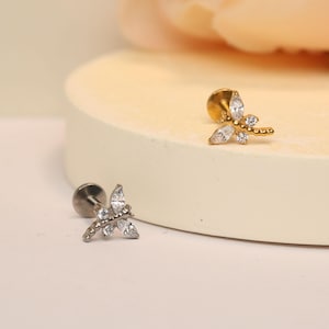 May include: Two silver and gold butterfly-shaped body jewelry pieces with clear crystal accents. The jewelry is on a white surface.