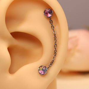 Puede incluir: Un piercing plateado con dos gemas redondas e iridiscentes conectadas por una delicada cadena. Las gemas muestran una mezcla de tonos rosas, morados y azules, creando un efecto sutil pero llamativo. La joya está diseñada para ser usada en la oreja.