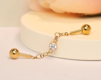 Op de afbeelding: Gouden kettingoorring met een heldere strasssteen in het midden. De oorring heeft twee gouden ballen aan beide uiteinden van de ketting.