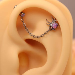 Puede incluir: Un piercing de oreja plateado con una cadena y dos elementos decorativos. Uno es una pequeña araña con una gema púrpura, el otro un pequeño perno redondo con una gema púrpura a juego. El piercing se muestra en una oreja de maniquí.