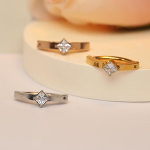 Puede incluir: Tres pendientes de aro con una piedra transparente de forma cuadrada en el centro. Los aros son de plata, oro y oro rosa.