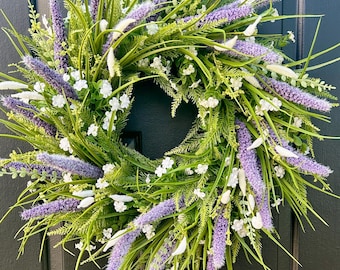Peut inclure: Une couronne de printemps avec des fleurs violettes et blanches et un feuillage vert. La couronne est faite de fleurs et de verdure artificielles.