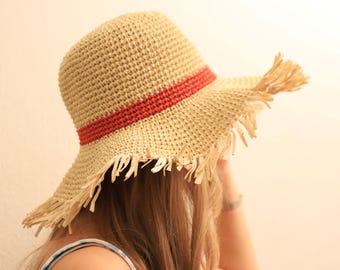 Puede incluir: Un sombrero de sol de paja con ala ancha y banda roja. El sombrero tiene un borde con flecos y está hecho de material de paja tejida. Un accesorio de verano para la protección solar.