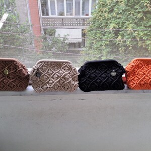 Op de afbeelding: Vier gehaakte tassen in verschillende kleuren: bruin, beige, zwart en oranje. De tassen zijn op een vensterbank uitgelijnd.