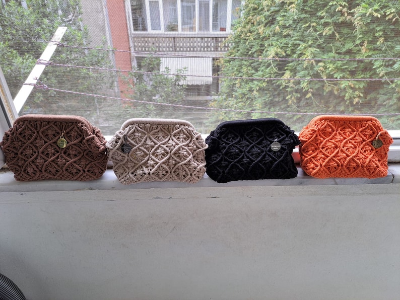 Op de afbeelding: Vier gehaakte tassen in verschillende kleuren: bruin, beige, zwart en oranje. De tassen zijn op een vensterbank uitgelijnd.