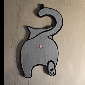 Puede incluir: Una figurita de gato gris con contorno negro, de pie sobre sus patas traseras con la cola enroscada sobre su espalda. El gato tiene una estrella rosa en su trasero y huellas de patas negras en su pata trasera.