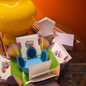 Może przedstawiać: Ręcznie robione pudełko eksplodujące z niebieskim wzorem postaci, zielonymi i fioletowymi akcentami oraz różnymi kartami dekoracyjnymi. Pudełko jest otwarte, ukazując wiele warstw i elementów pop-up. W tle żółty wazon i pomarańczowa ściana.