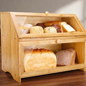 May include: A two-tier bamboo bread box with clear glass windows on the front panels. The top tier contains assorted muffins and sliced bread, while the bottom tier holds a loaf of bread and several slices of dark bread, all accessible via knobs.