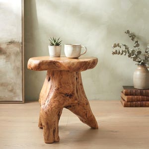 May include: A natural light brown wooden stool crafted from a single tree stump with a thick, uneven circular top and three sturdy, organic-shaped legs. The surface of the wood displays visible grain patterns, knots, and a smooth, polished finish.