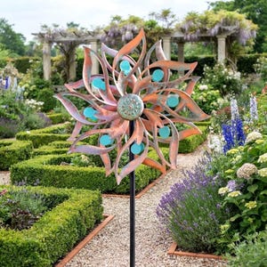 May include: A metal wind spinner featuring a multi-layered, flower-like design with copper-toned petals and small, circular turquoise accents. The center is a textured, round medallion, and the entire structure is mounted on a thin black pole.