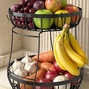 May include: A two-tiered black metal wire basket stand filled with fresh produce. The top tier holds red cherries, purple plums, green apples, and yellow citrus. The bottom tier contains garlic bulbs, sweet potatoes, red onions, and yellow bananas.