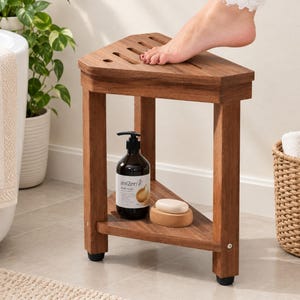 May include: A triangular brown wooden shower stool with a slatted top surface and a lower storage shelf. The stool features four sturdy legs with black rubber feet. A dark bottle of LexiZen body wash and a bar of soap sit on the lower shelf.