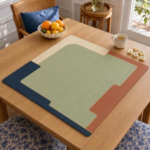 Può includere: Un tappetino da gioco quadrato e multicolore caratterizzato da un ampio pannello centrale verde salvia con superficie testurizzata, bordato da sezioni color crema, blu navy e arancione terracotta. Il tappetino è appoggiato su un tavolo in legno.