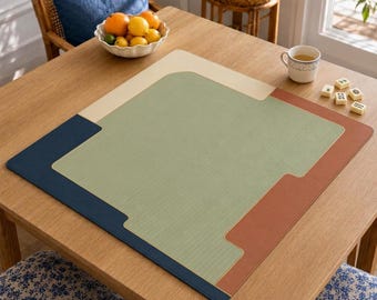 May include: A square, multi-colored game mat featuring a large central sage green panel with a textured surface, bordered by sections in cream, navy blue, and terracotta orange. The mat lies flat on a wooden table surface with a smooth, clean finish.