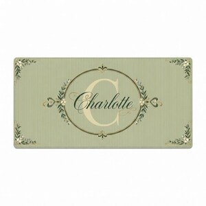 May include: A rectangular sage green mat featuring a thin vertical pinstripe pattern. In the centre, a large cream-coloured letter C is overlaid with the name Charlotte in dark green script, enclosed within an oval frame with floral accents.