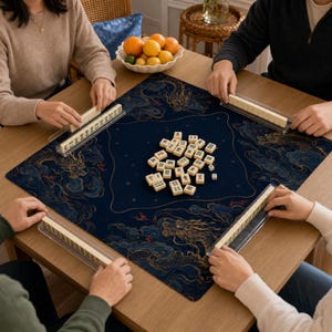 May include: A square mahjong mat featuring a dark blue background with gold and blue dragon and cloud patterns. The mat is placed on a wooden table with a central pile of ivory-colored mahjong tiles and four clear plastic tile racks.