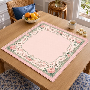 May include: A square table mat featuring a solid light pink center framed by a decorative border of green foliage, pink peony flowers, and small birds. The mat has a scalloped inner edge design and a thin pink outer trim on a wooden table.