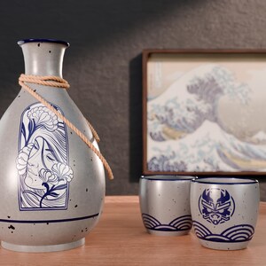 May include: A gray ceramic sake set with a floral design on the bottle and a wave design on the cups. The set includes a bottle, two cups, and a rope.