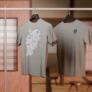 May include: Two gray t-shirts hanging on a rack. One t-shirt has a graphic of a stylized face with leaves. The other t-shirt has a small graphic of a stylized bird.