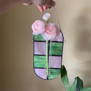 May include: A stained glass bud vase with a chain for hanging, holding pink flowers. The vase has a green and pink striped design. The flowers are light pink and the vase is held by a silver chain.