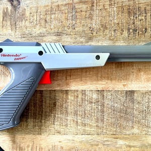 May include: A grey Nintendo Zapper light gun controller featuring a bright red trigger, a ribbed handle grip, and a long barrel. The side of the device is clearly marked with the text Nintendo Zapper and the year 1985, with a black cord extending from the base.