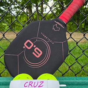 Puede incluir: Una pala de pickleball negra y rosa con el texto "O-G" en rosa. Dos pelotas de pickleball amarillas con una banda blanca que dice "CRUZ" están delante de la pala.