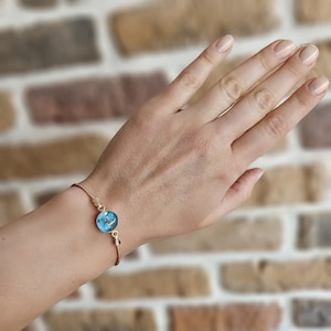 May include: A gold-coloured bracelet with a round, blue pendant featuring a swirling design. The bracelet has a fine chain and a small clasp. The pendant's design includes shades of blue and white.