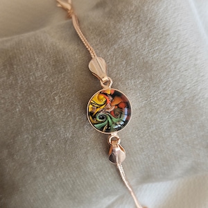 May include: A gold chain bracelet with a round, colorful glass pendant. The pendant features a swirling pattern of green, yellow, orange, and black.