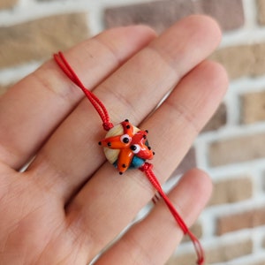 Op de afbeelding: Rood koord armband met een kleurrijke glazen zeester bedel. De zeester heeft oranje, rode, witte en blauwe details.