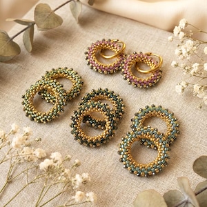 Pode incluir: Uma variedade de brincos de argola com contas em várias cores, incluindo dourado, verde e rosa. Os brincos estão dispostos sobre uma superfície de tons neutros, com raminhos de vegetação e flores brancas que adicionam um toque natural. Os brincos têm um design circular.