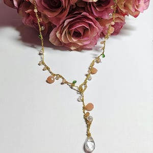 May include: A gold chain necklace with a teardrop-shaped crystal pendant. The necklace is adorned with small pink, white, and green beads.