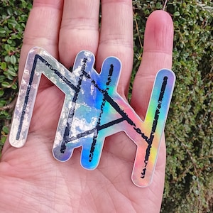 Può includere: Un adesivo olografico a forma di runa, con linee nere e un bagliore arcobaleno. L'adesivo è tenuto in una mano su uno sfondo di fogliame verde.